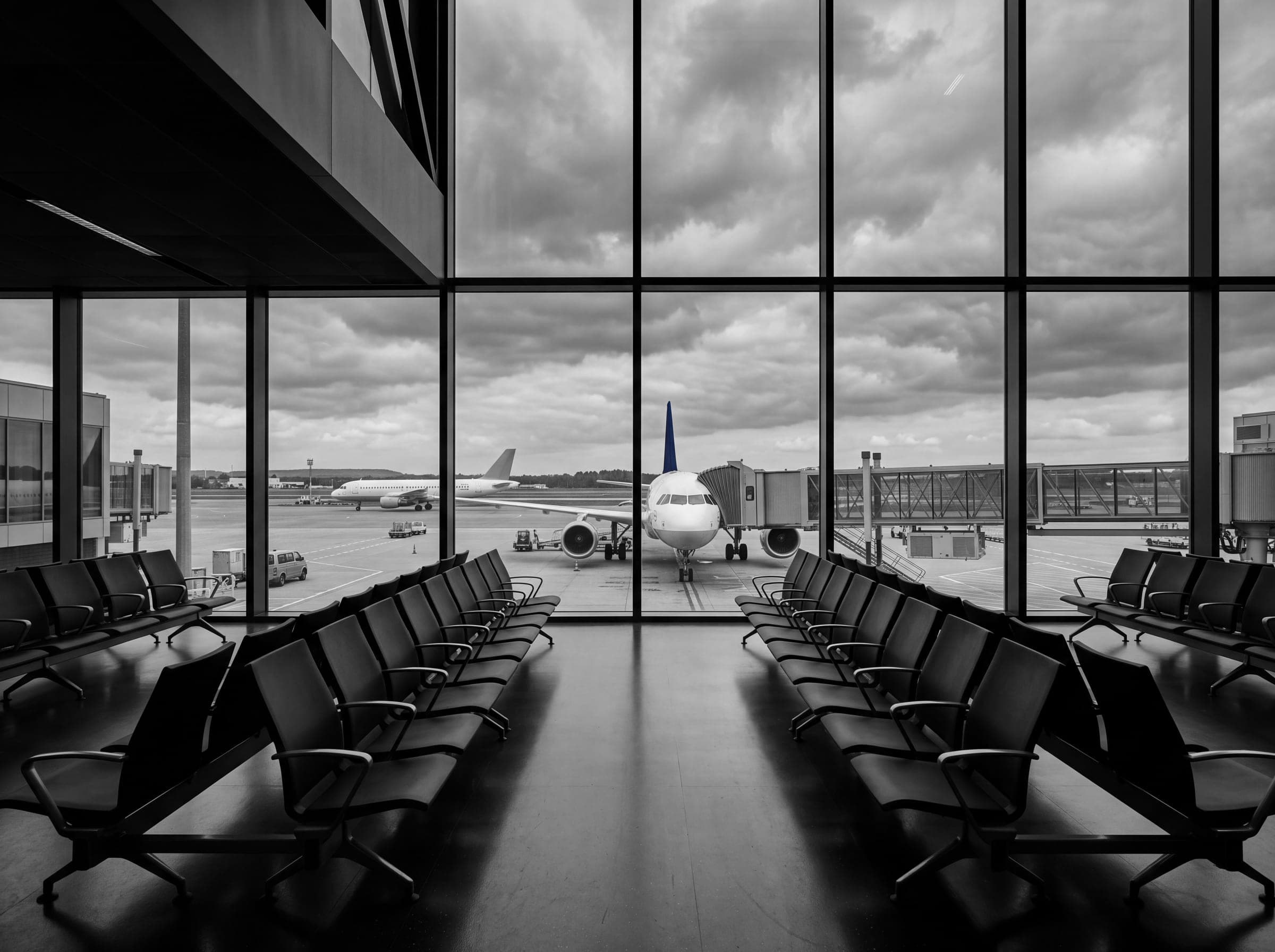 An empty departure gate in black and white: rows of seats leading to tall windows, and beyond them a passenger jet at the gate with its tail in muted navy.